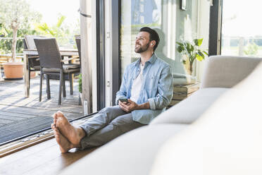 Thoughtful freelancer wearing wireless in-ear headphones sitting with smart phone at home - UUF29099