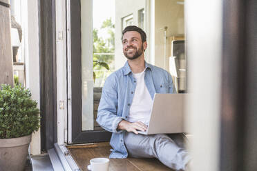 Happy businessman sitting with laptop in doorway - UUF29108