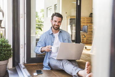 Glücklicher Geschäftsmann sitzt mit Kaffeetasse und Laptop auf dem Boden in der Tür - UUF29110