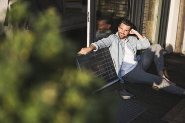 Lächelnder Geschäftsmann sitzt mit Solarpanel auf der Veranda - UUF29122