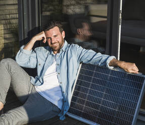 Glücklicher Geschäftsmann sitzt mit Solarpanel auf der Veranda - UUF29123