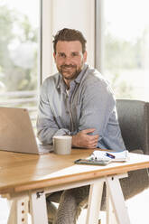 Happy businessman sitting at desk in home office - UUF29130