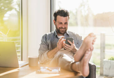 Smiling businessman using smart phone in home office - UUF29137