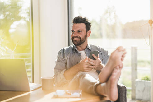 Happy businessman sitting with mobile phone in home office - UUF29138