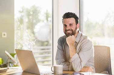 Happy businessman with hand on chin sitting at desk in home office - UUF29141