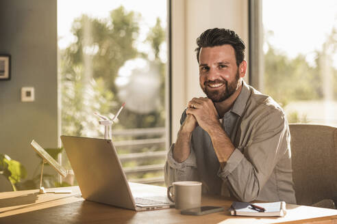 Glücklicher Geschäftsmann sitzt mit Laptop am Schreibtisch im Heimbüro - UUF29142