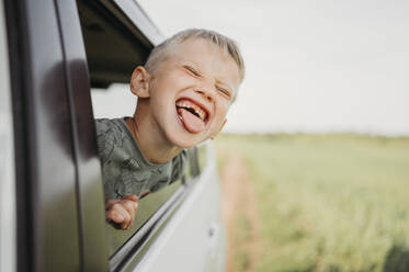 Lächelnder Junge mit herausgestreckter Zunge, der aus dem Autofenster schaut - ANAF01554