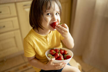 Smiling boy eating strawberry from bowl at home - ANAF01569