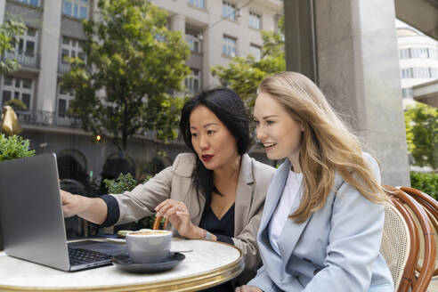 Businesswoman showing over laptop to partner at sidewalk cafe - NDEF00839