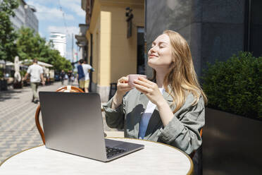 Geschäftsfrau mit geschlossenen Augen genießt einen Kaffee vor einem Laptop in einem Straßencafé - NDEF00858