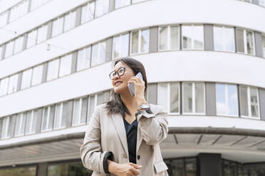 Lächelnde Geschäftsfrau, die vor einem Gebäude steht und mit einem Smartphone spricht - NDEF00868