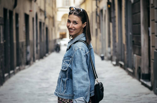 Smiling woman looking over shoulder standing in alley - JSRF02549