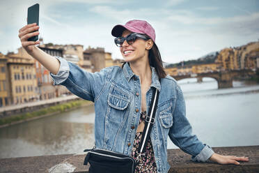 Eine fröhliche Frau hält einen Moment auf einer Brücke mit ihrem Smartphone fest und lächelt für ein Selfie - JSRF02554