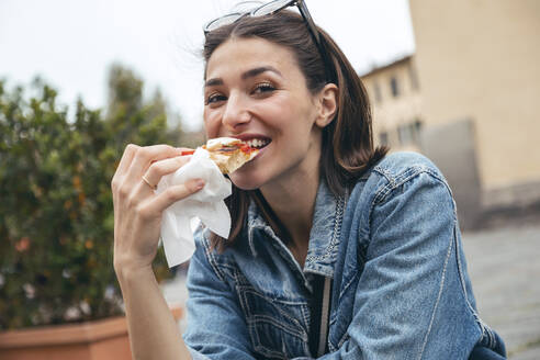 Smiling woman eating pizza sitting on road - JSRF02562