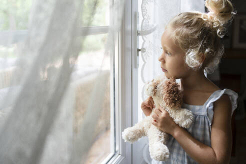 Nachdenkliches Mädchen, das ein Kuscheltier hält und aus dem Fenster schaut, zu Hause - SVKF01474