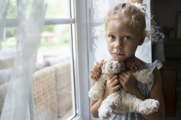 Sad girl holding stuffed toy standing near window at home - SVKF01475