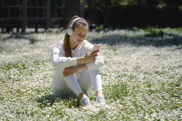 Teenager-Mädchen genießt Musik mit Kopfhörern auf einem Blumenbeet im Park sitzend - AAZF00758