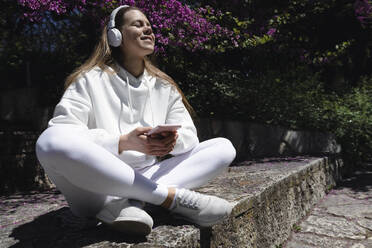 Teenage girl listening music with headphones on wall at park - AAZF00761