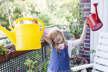 Smiling girl standing with watering cans in balcony - IHF01411