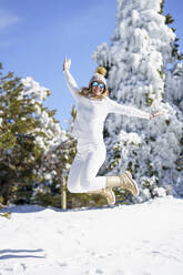 Smiling woman wearing white outfit jumping on winter snow - JSMF02786
