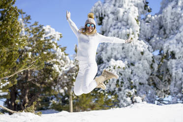 Lächelnde Frau mit weißem Outfit und Sonnenbrille springt auf Schnee - JSMF02787