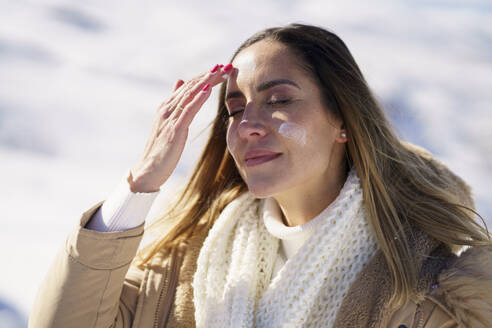 Lächelnde Frau mit geschlossenen Augen, die sich im Winter mit Sonnencreme eincremt - JSMF02804