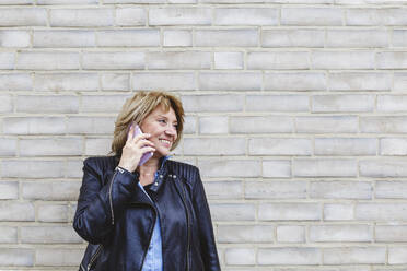 Senior woman talking on smart phone in front of brick wall - IHF01448