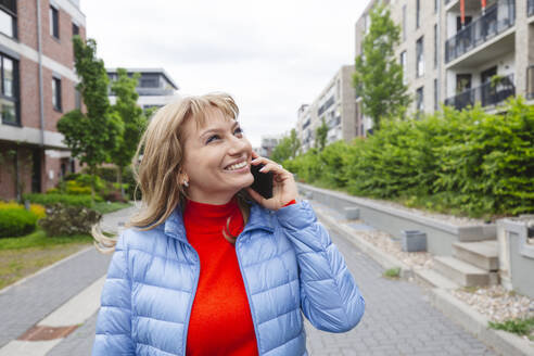 Lächelnde Frau, die in der Nähe eines Wohngebiets mit einem Smartphone telefoniert - IHF01459