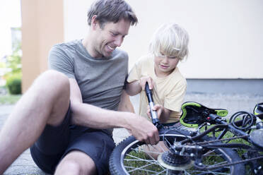 Glücklicher Vater und Sohn mit Pumpe reparieren Fahrrad am Fußweg - NJAF00427