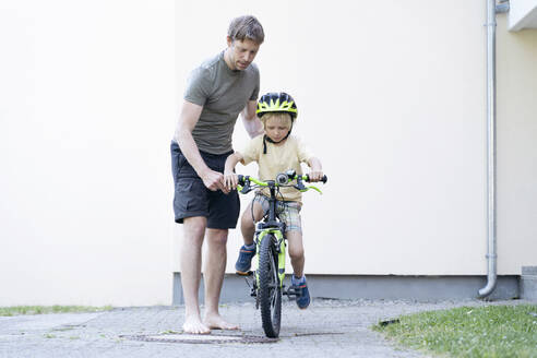 Vater mit Sohn beim Fahrradfahren vor einem Gebäude - NJAF00431