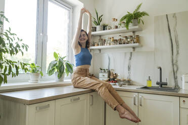 Glückliche junge Frau, die ihre Arme auf dem Küchentisch zu Hause ausstreckt - OSF01777