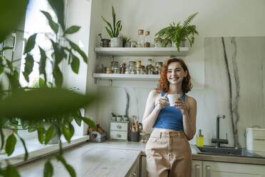 Happy young woman standing with coffee cup in kitchen at home - OSF01779
