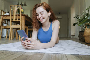 Glückliche junge Frau benutzt ihr Smartphone auf dem Teppich zu Hause - OSF01797