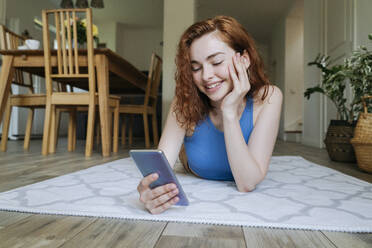 Glückliche junge Frau mit Hand am Kinn, die zu Hause ein Smartphone benutzt - OSF01798