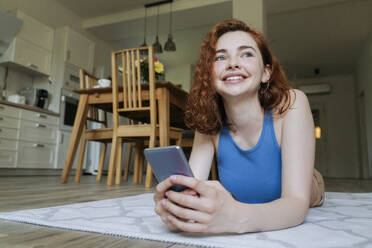 Glückliche junge Frau mit Smartphone, die zu Hause auf dem Teppich liegt - OSF01799