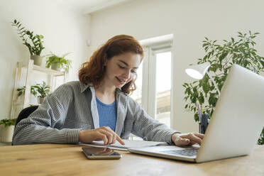 Happy freelancer with document and laptop sitting at table - OSF01814