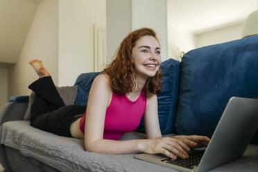 Happy young woman lying on sofa with laptop at home - OSF01823