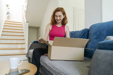 Glückliche junge Frau beim Auspacken eines Pakets auf dem Sofa zu Hause - OSF01826