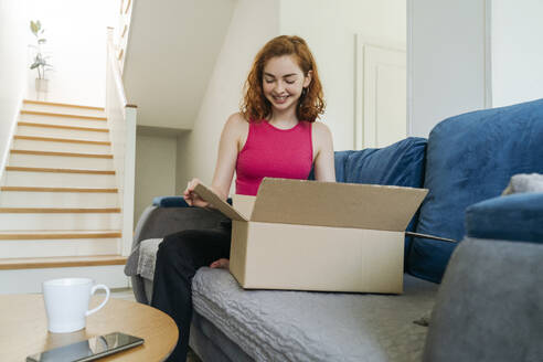 Happy young woman unboxing package on sofa at home - OSF01826