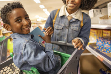 Lächelnder Junge im Einkaufswagen mit Mutter im Supermarkt - JCCMF10500