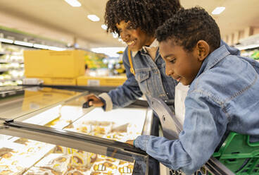 Glückliche Mutter und Sohn betrachten Tiefkühlkost in der Gefriertruhe im Supermarkt - JCCMF10504