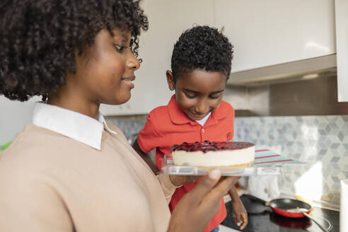 Lächelnder Junge mit Mutter, die Kuchen in der Küche zu Hause hält - JCCMF10528