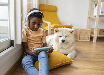 Boy wearing wireless headphones using mobile phone and sitting by dog at home - JCCMF10562