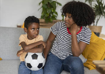 Sad boy holding soccer ball and sitting with mother on sofa at home - JCCMF10575