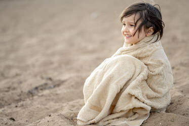 Happy boy wrapped in towel sitting on sand at beach - ANAF01591