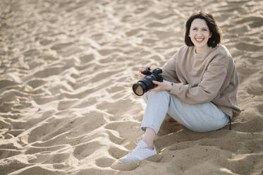 Glückliche Frau sitzt mit Kamera auf Sand am Strand - ANAF01593
