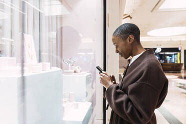 Smiling woman using smart phone standing in front of store window - PNAF05526