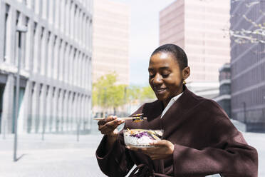 Lächelnde Geschäftsfrau mit Essstäbchen beim Essen im Büropark - PNAF05539