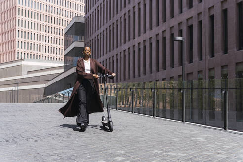 Geschäftsfrau zu Fuß mit Elektro-Scooter vor einem Gebäude - PNAF05549