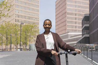 Happy young businesswoman standing with electric push scooter on sunny day - PNAF05552